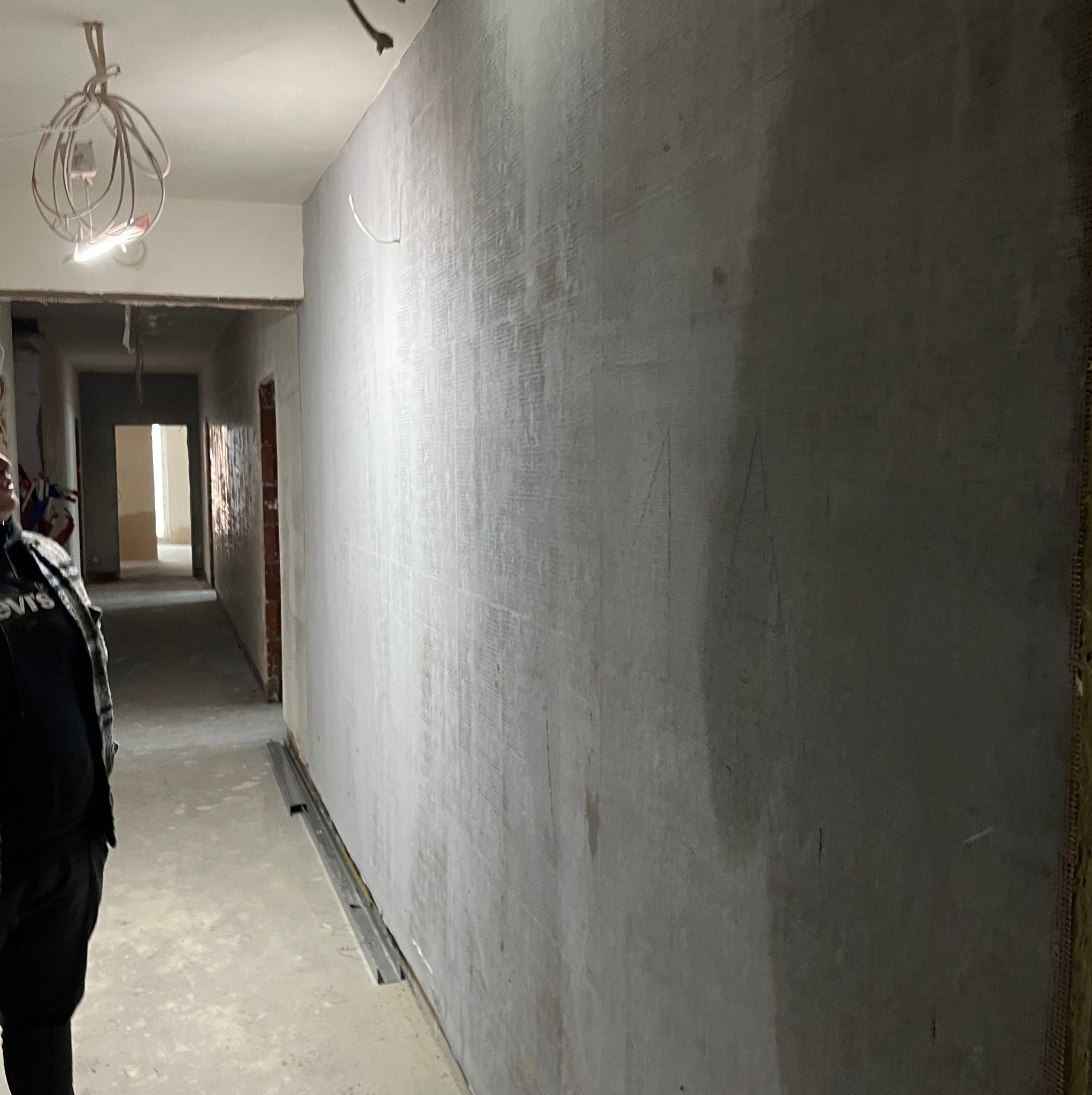 Ściana w trakcie szpachlowania z widoczną siatką wzmacniającą, surowy beton na podłodze, widok w głąb korytarza z otwartymi drzwiami, prowizoryczne oświetlenie zwisające z sufitu.