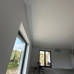 MR. WHITE Marek Ryborz - Wnętrze po remoncie: białe ściany, sufit i nietypowe okno trójkątne. Na podłodze widoczne ślady po pracach. W tle drzwi balkonowe i krzesło.