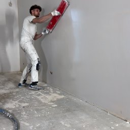 MR. WHITE Marek Ryborz - Pracownik w białym kombinezonie wyrównuje ścianę czerwoną szpachlą. Widoczny fragment podłogi z resztkami materiałów budowlanych i wąż od urządzenia.