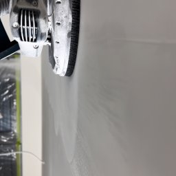 MR. WHITE Marek Ryborz - Szlifierka do gładzi w trakcie pracy na ścianie, widoczne ślady szlifowania. Ujęcie z bliska, skupione na detalu narzędzia i powierzchni ściany.