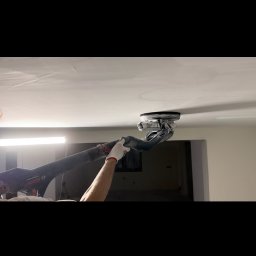 MR. WHITE Marek Ryborz - Szlifowanie sufitu gipsowego za pomocą szlifierki teleskopowej Bosch. Pracownik w rękawicach. Widoczne ślady po szlifowaniu. W tle ściana i oświetlenie.