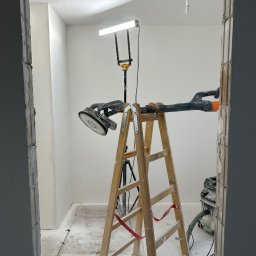 MR. WHITE Marek Ryborz - Pokój w trakcie prac wykończeniowych: drabina, szlifierka do gipsu z odkurzaczem, lampa oświetleniowa. Widok przez surową futrynę. Ściany przygotowane do malowania.