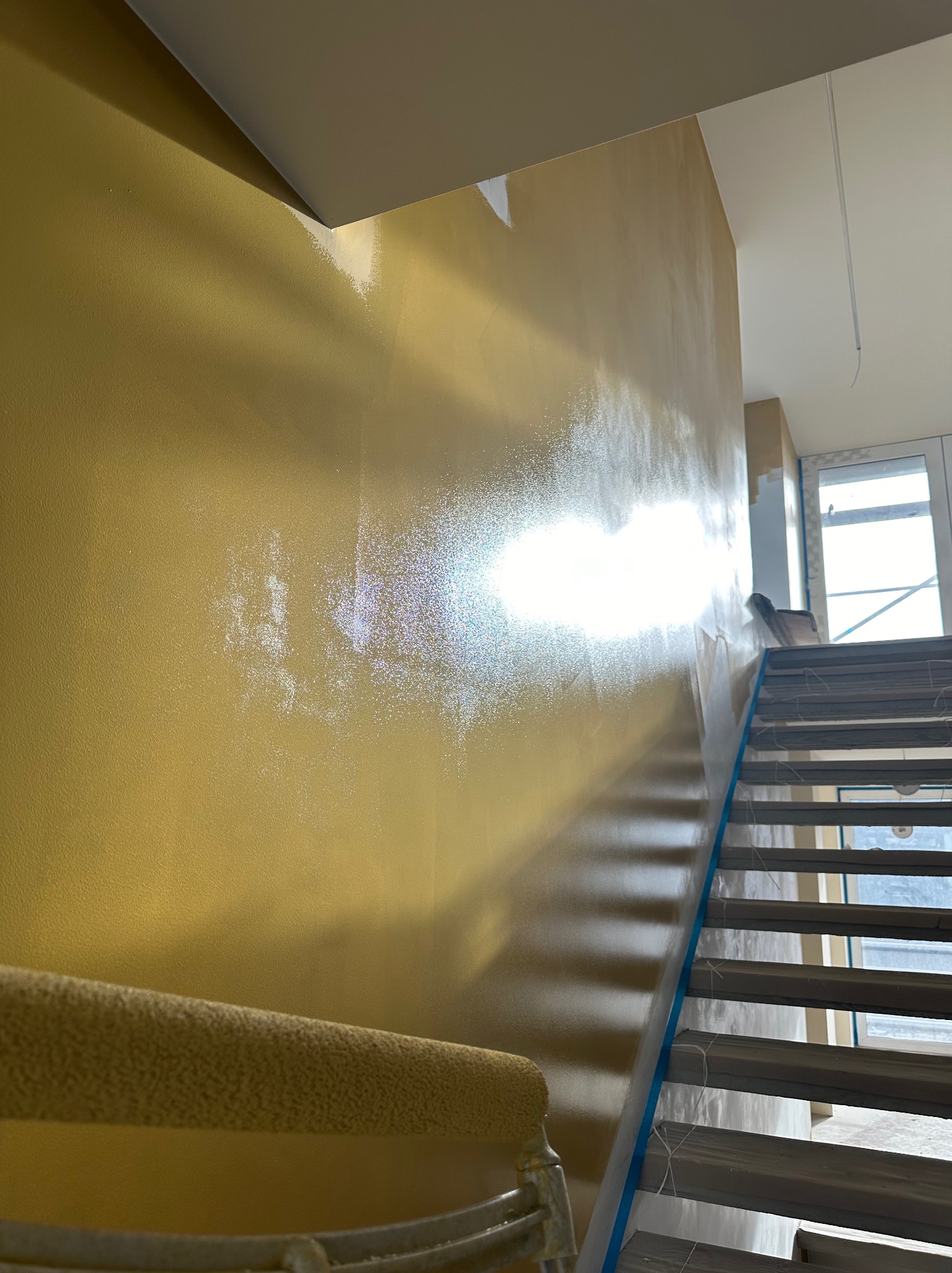 Ściana w trakcie malowania na złoty kolor, widoczne schody zabezpieczone folią i wałek malarski. Częściowo pomalowana ściana w klatce schodowej.