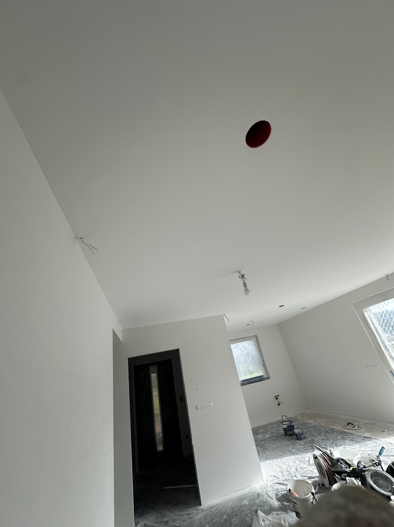 Wnętrze po malowaniu na biało, widoczne gładkie ściany i sufit z otworem na oświetlenie. Okno z widokiem na zewnątrz. Podłoga zabezpieczona folią malarską.