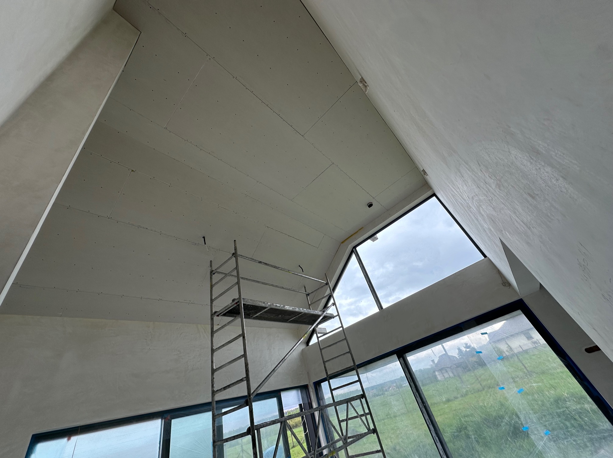 Wysokie wnętrze w trakcie wykańczania: sufit z płyt gipsowo-kartonowych, ściany po szpachlowaniu, rusztowanie na tle okien z widokiem na zieleń. Nietypowy kształt pomieszczenia.