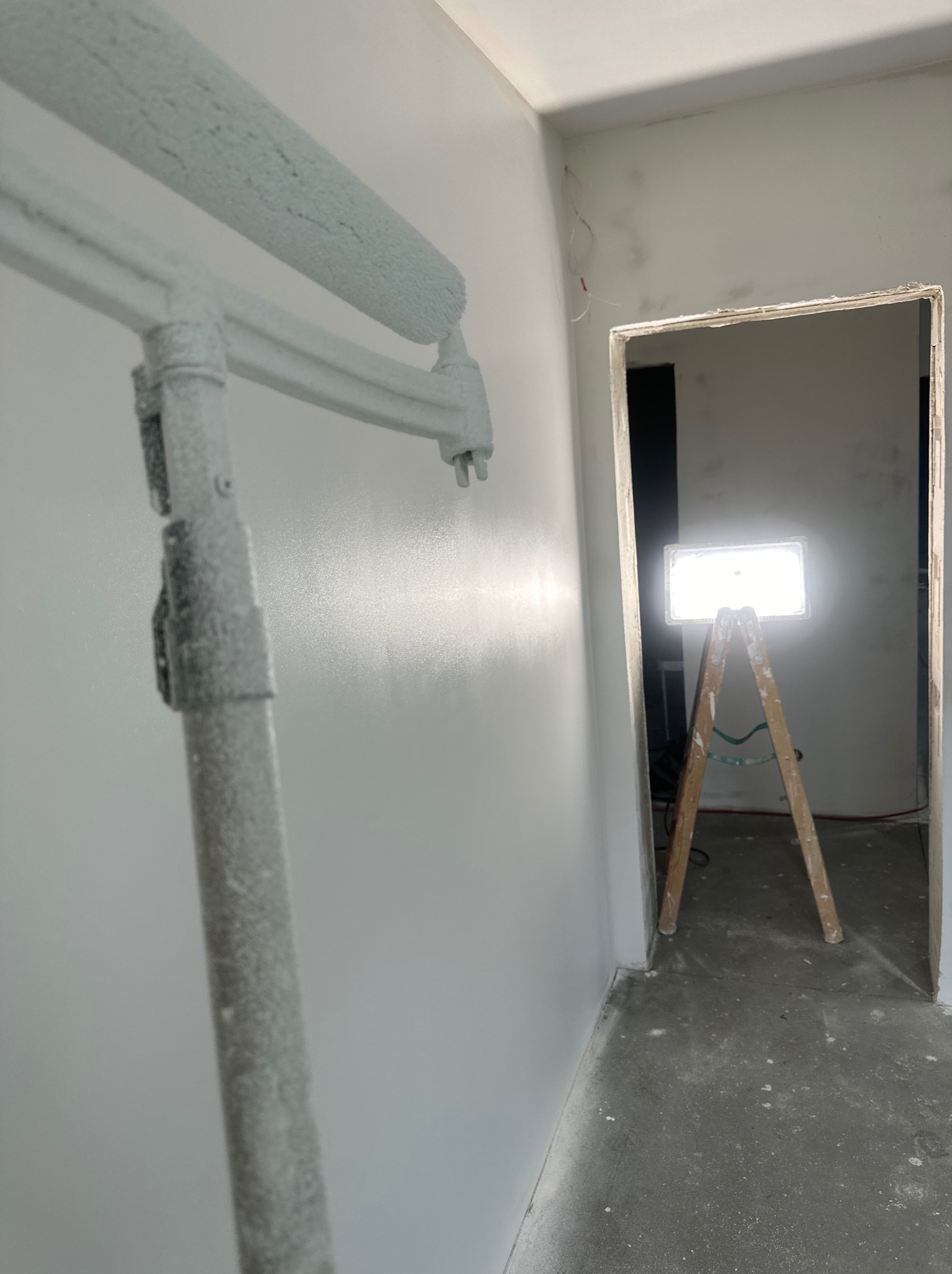 Świeżo pomalowana ściana z wałkiem malarskim w kadrze, w tle drabina z lampą oświetlającą wnętrze. Widoczne niedoskonałości na ścianie przed malowaniem.