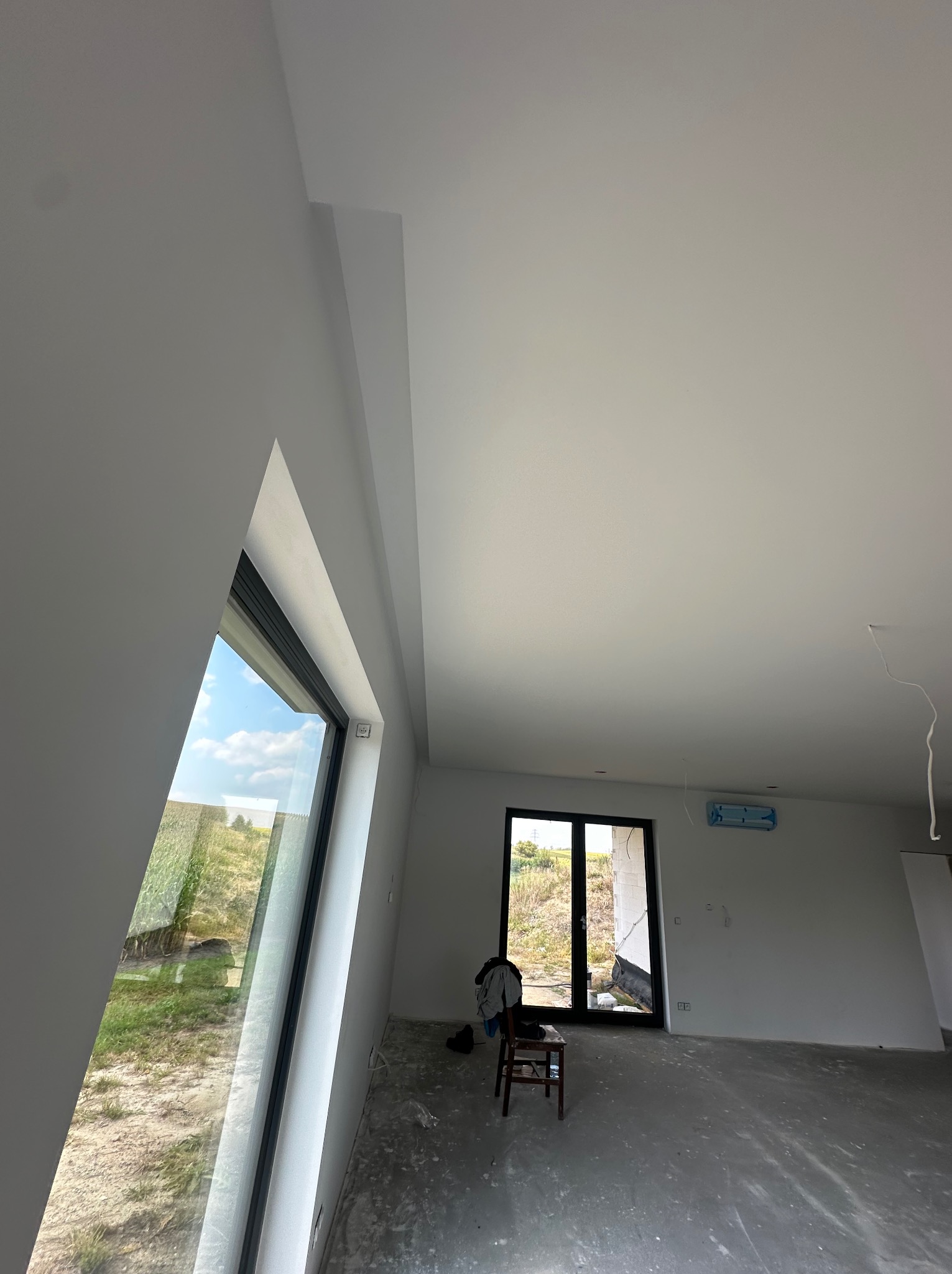 Wnętrze po remoncie: białe ściany, sufit i nietypowe okno trójkątne. Na podłodze widoczne ślady po pracach. W tle drzwi balkonowe i krzesło.