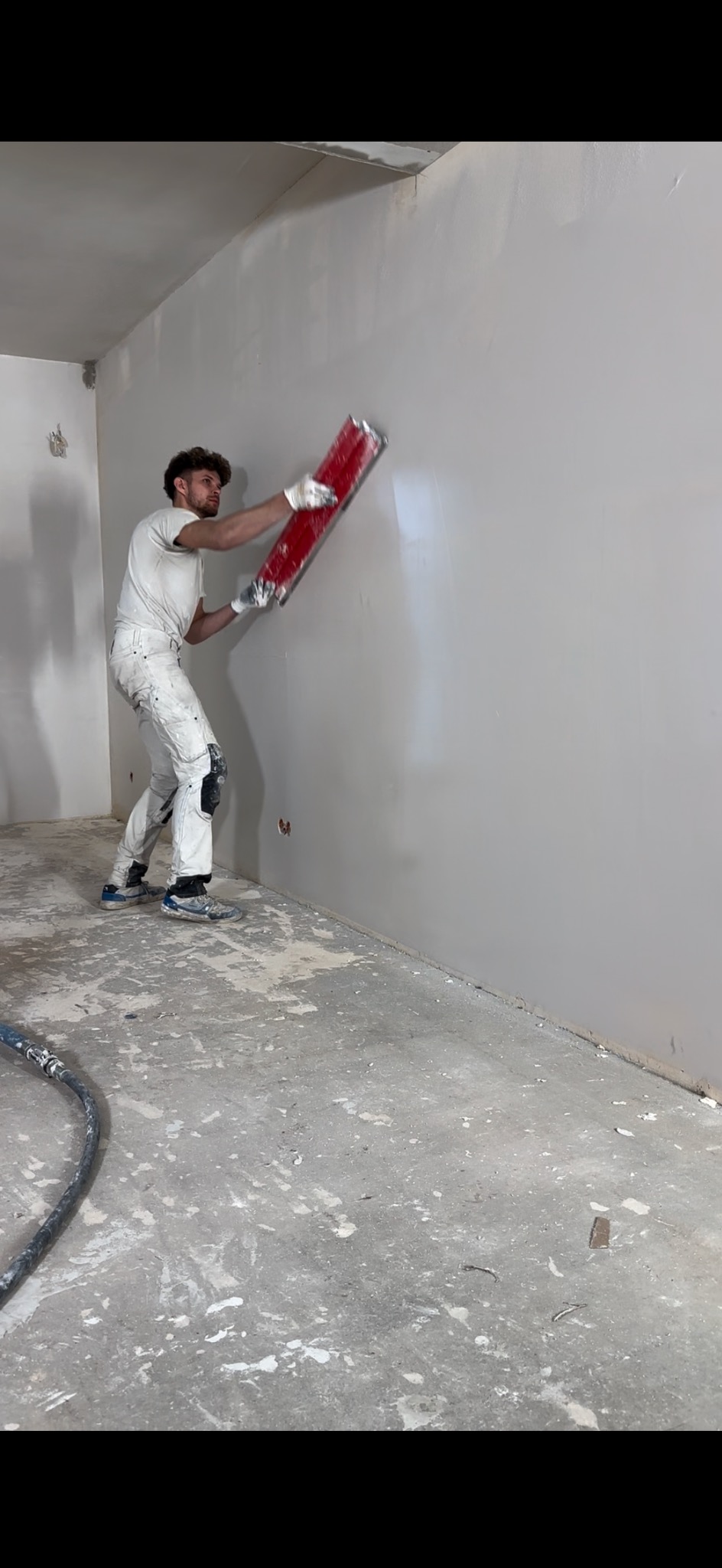 Pracownik w białym kombinezonie wyrównuje ścianę czerwoną szpachlą. Widoczny fragment podłogi z resztkami materiałów budowlanych i wąż od urządzenia.
