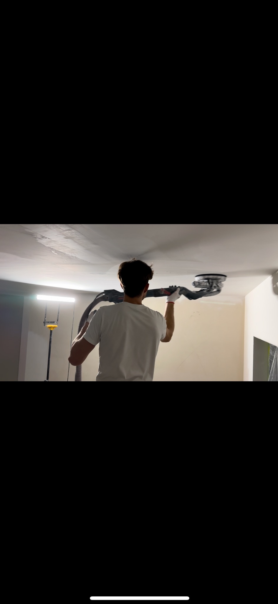Pracownik w białej koszulce szlifuje sufit za pomocą szlifierki z odkurzaczem. Widoczne ślady po szpachlowaniu i oświetlenie robocze.