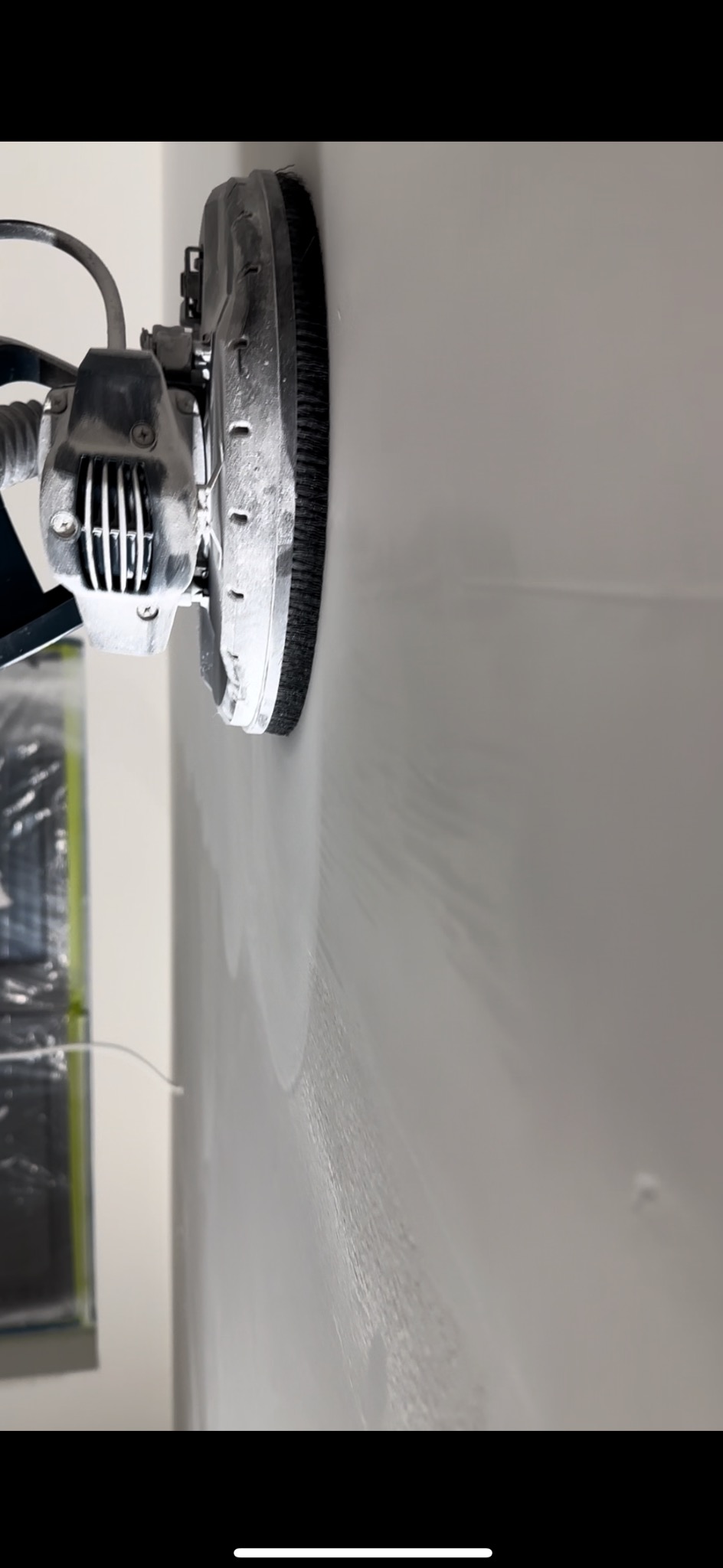 Szlifierka do gładzi w trakcie pracy na ścianie, widoczne ślady szlifowania. Ujęcie z bliska, skupione na detalu narzędzia i powierzchni ściany.