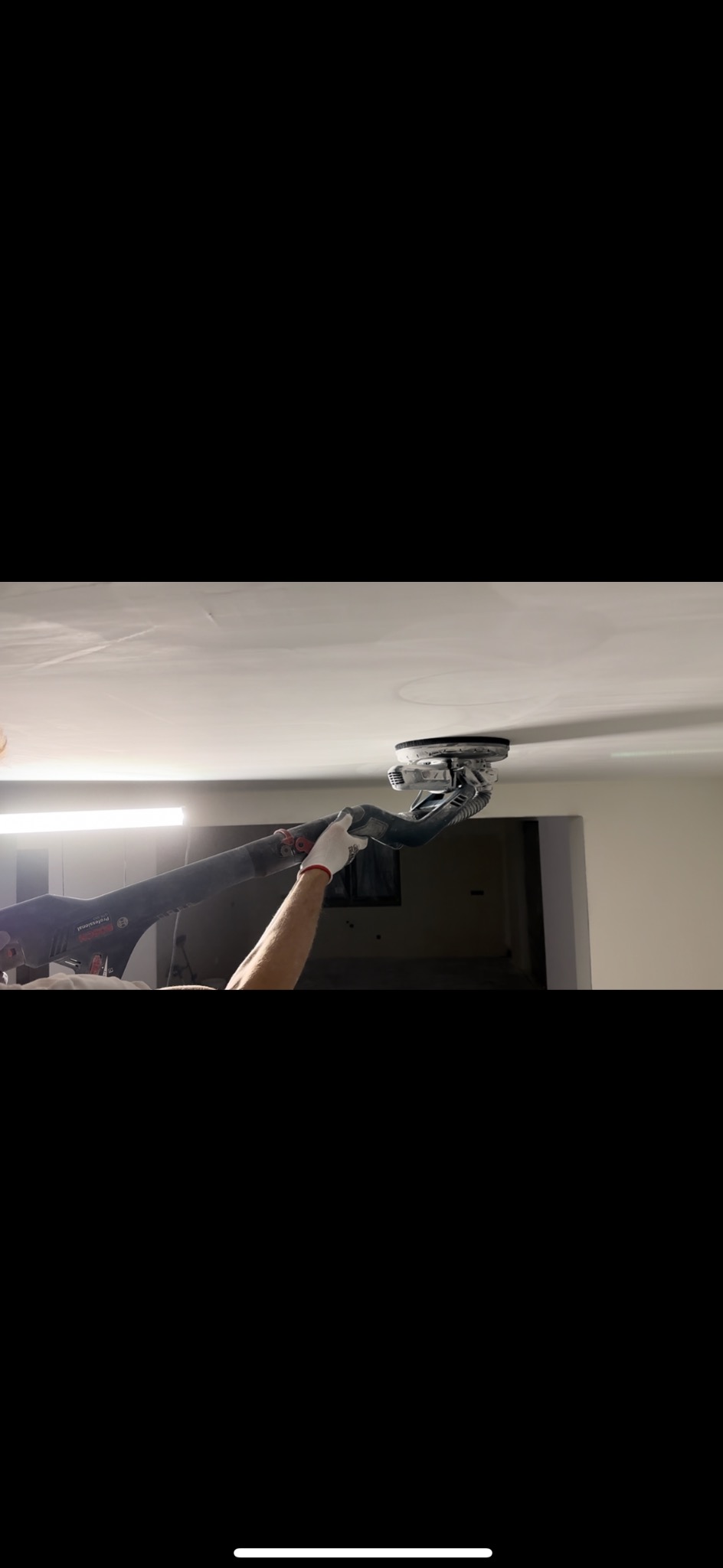 Szlifowanie sufitu gipsowego za pomocą szlifierki teleskopowej Bosch. Pracownik w rękawicach. Widoczne ślady po szlifowaniu. W tle ściana i oświetlenie.