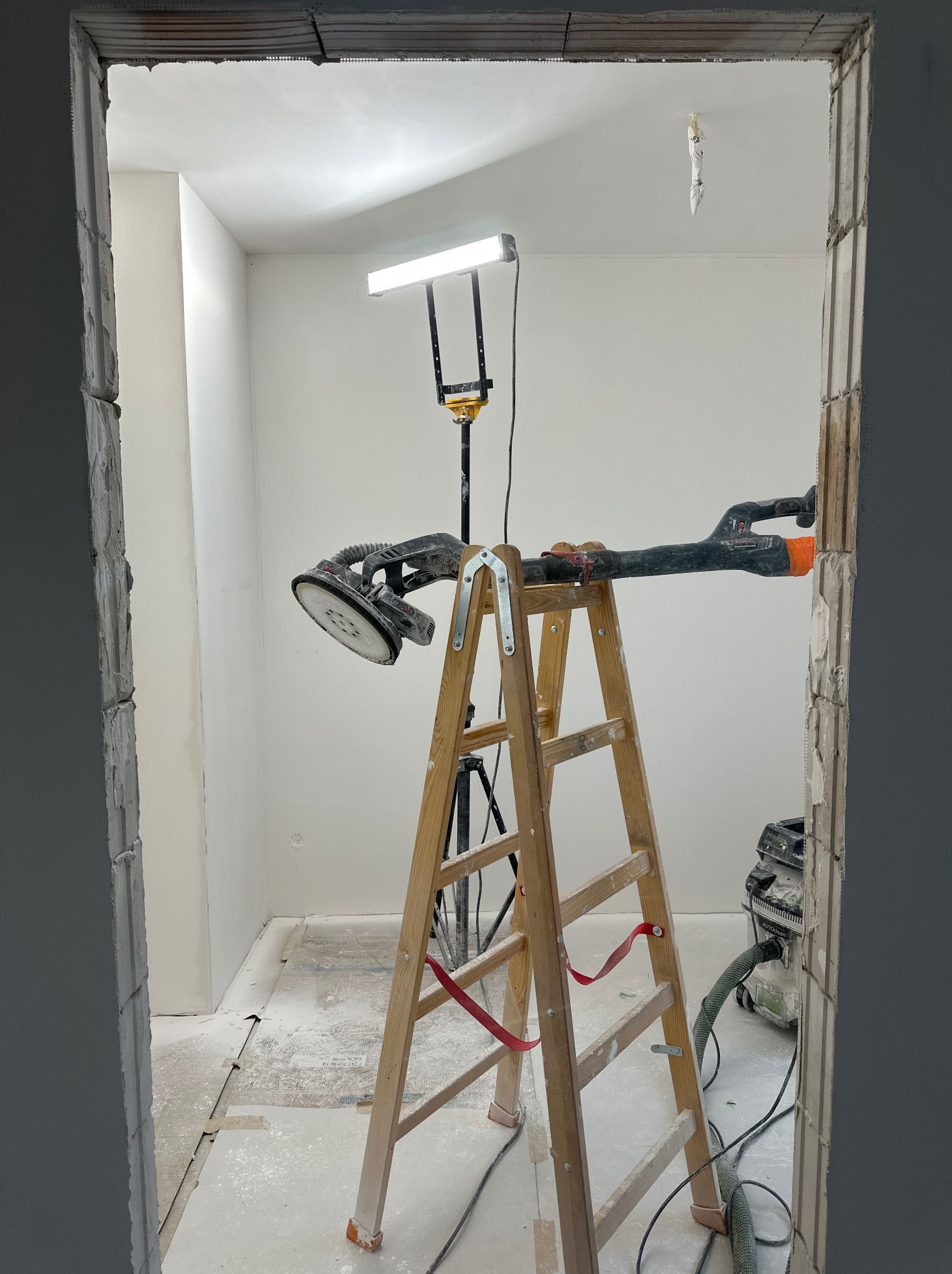 Pokój w trakcie prac wykończeniowych: drabina, szlifierka do gipsu z odkurzaczem, lampa oświetleniowa. Widok przez surową futrynę. Ściany przygotowane do malowania.