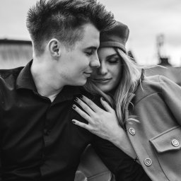 Jakub Matuszny jamafoto.pl - Czarno-białe zdjęcie zakochanej pary: mężczyzna w czarnej koszuli przytula kobietę w płaszczu i berecie, na jej palcu widoczny pierścionek zaręczynowy.