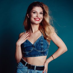 Jakub Matuszny jamafoto.pl - Portret uśmiechniętej blondynki w dżinsowym topie i spodniach, na jednolitym turkusowym tle, z widocznym złotym bransoletką i czerwonymi ustami.