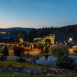 Jakub Matuszny jamafoto.pl - Wieczorny widok na kompleks budynków z panelami słonecznymi, oświetlony dekoracyjnymi lampkami, z sauną beczką i oczkiem wodnym na pierwszym planie.
