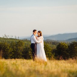 Jakub Matuszny jamafoto.pl - Para młoda w objęciach na tle malowniczego krajobrazu górskiego, ona w koronkowej sukni z welonem, on w białej koszuli i szelkach, złote trawy na pierwszym planie.