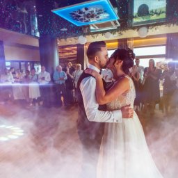 Jakub Matuszny jamafoto.pl - Pocałunek pary młodej podczas pierwszego tańca, otoczonej gośćmi w sali balowej z dekoracyjnym oświetleniem i efektem dymu.