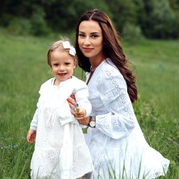 Jakub Matuszny jamafoto.pl - Uśmiechnięta matka z córką w identycznych białych, ażurowych sukienkach, siedząca na łące pełnej trawy i drobnych niebieskich kwiatów, trzymająca mały bukiecik.