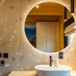 Jakub Matuszny jamafoto.pl - Nowoczesna łazienka z okrągłym lustrem podświetlanym LED, białą umywalką nablatową, czarną armaturą i ścianami wykończonymi płytkami imitującymi lastryko. W odbiciu lustra widoczna drewniana ściana...