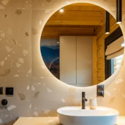 Nowoczesna łazienka z okrągłym lustrem podświetlanym LED, białą umywalką nablatową, czarną armaturą i ścianami wykończonymi płytkami imitującymi lastryko. W odbiciu lustra widoczna drewniana ściana...