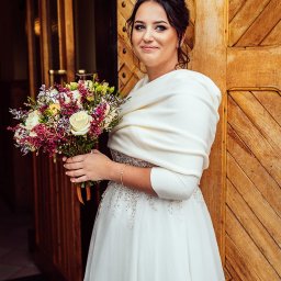 Jakub Matuszny jamafoto.pl - Uśmiechnięta panna młoda w białej sukni z delikatnymi zdobieniami i kremowym szalem, trzymająca bukiet z białych róż i fioletowych kwiatów na tle drewnianych drzwi.