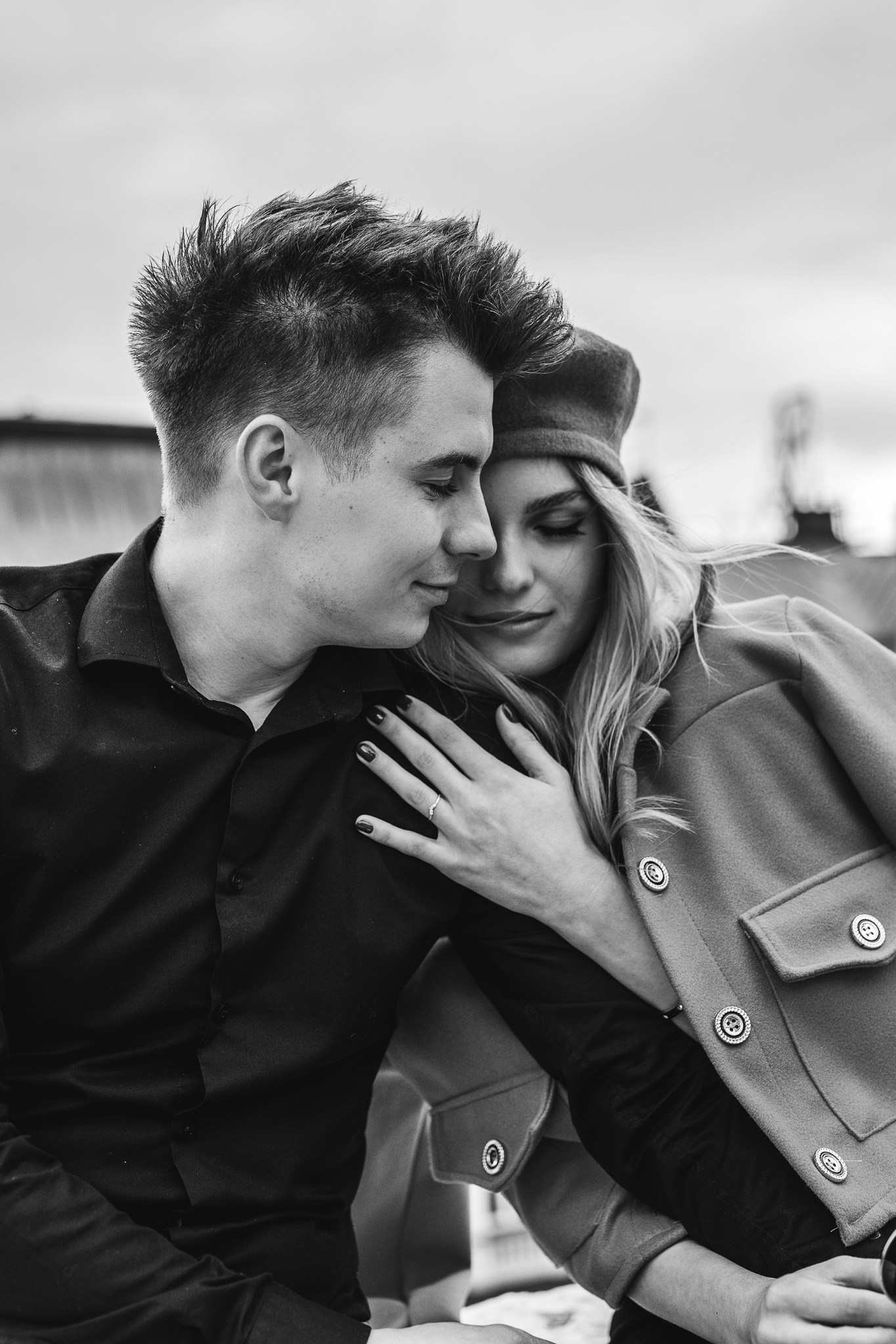 Czarno-białe zdjęcie zakochanej pary: mężczyzna w czarnej koszuli przytula kobietę w płaszczu i berecie, na jej palcu widoczny pierścionek zaręczynowy.