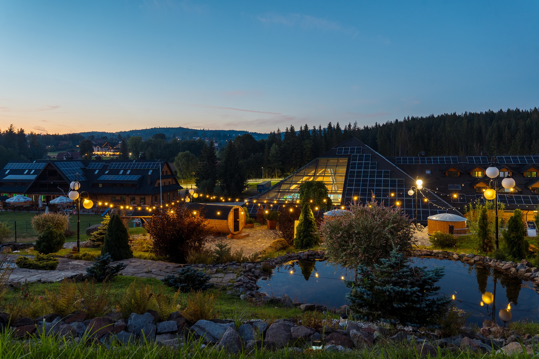 Wieczorny widok na kompleks budynków z panelami słonecznymi, oświetlony dekoracyjnymi lampkami, z sauną beczką i oczkiem wodnym na pierwszym planie.