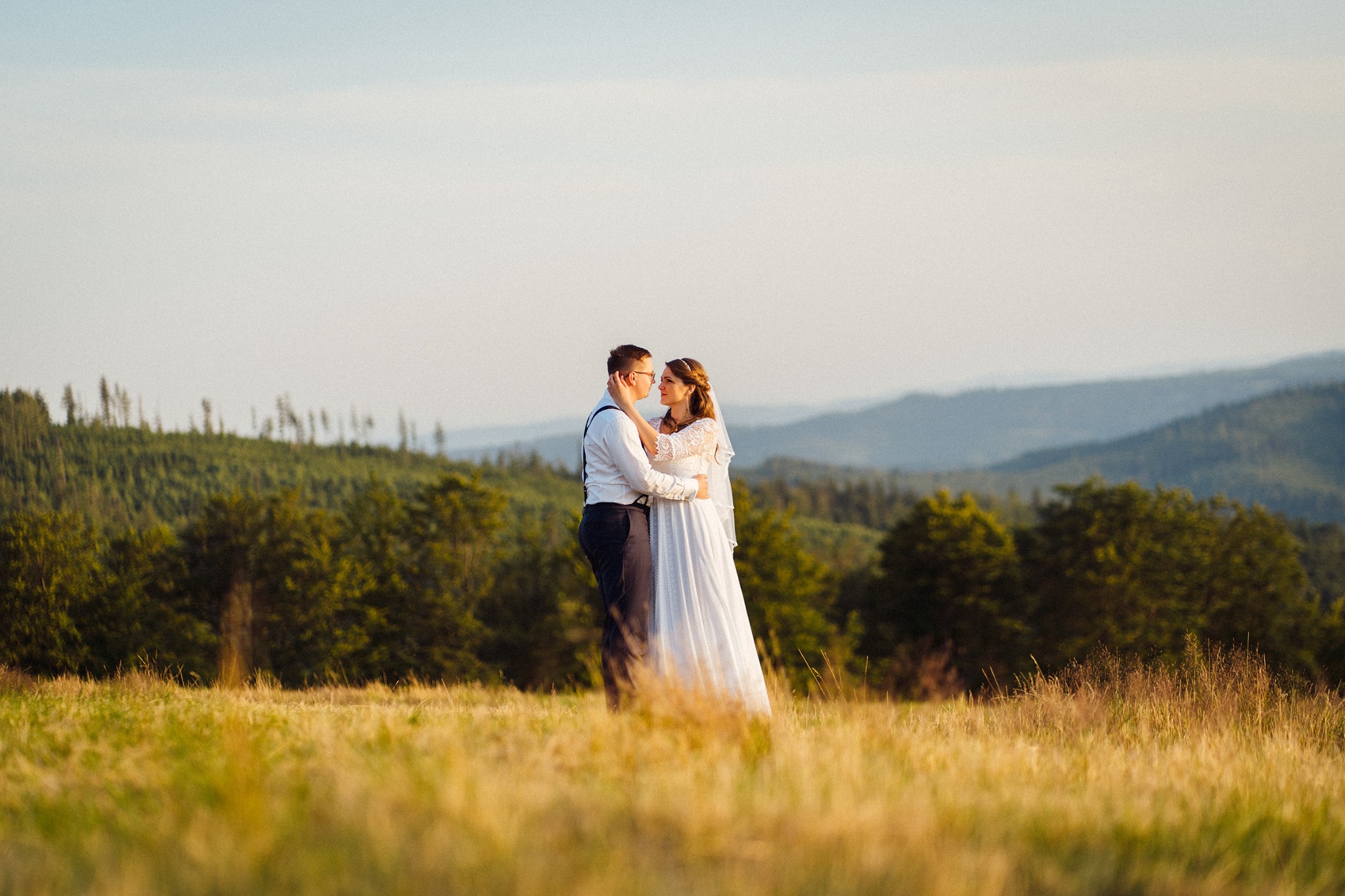 Para młoda w objęciach na tle malowniczego krajobrazu górskiego, ona w koronkowej sukni z welonem, on w białej koszuli i szelkach, złote trawy na pierwszym planie.