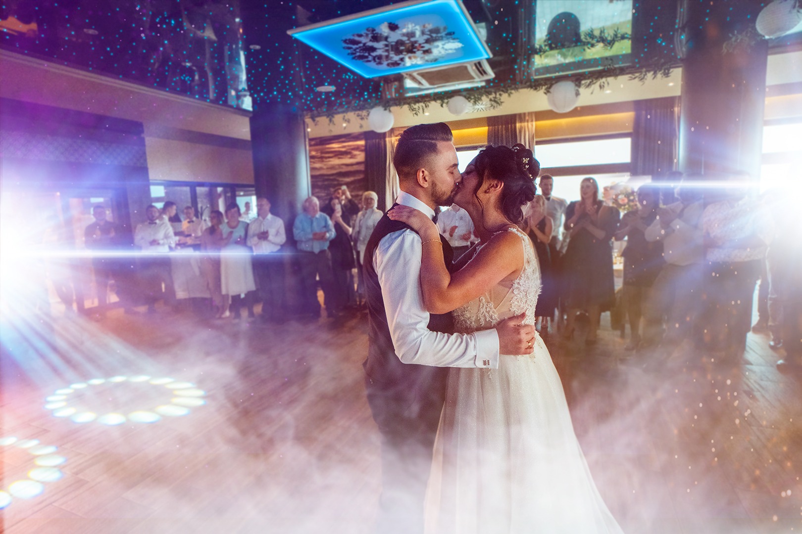 Pocałunek pary młodej podczas pierwszego tańca, otoczonej gośćmi w sali balowej z dekoracyjnym oświetleniem i efektem dymu.