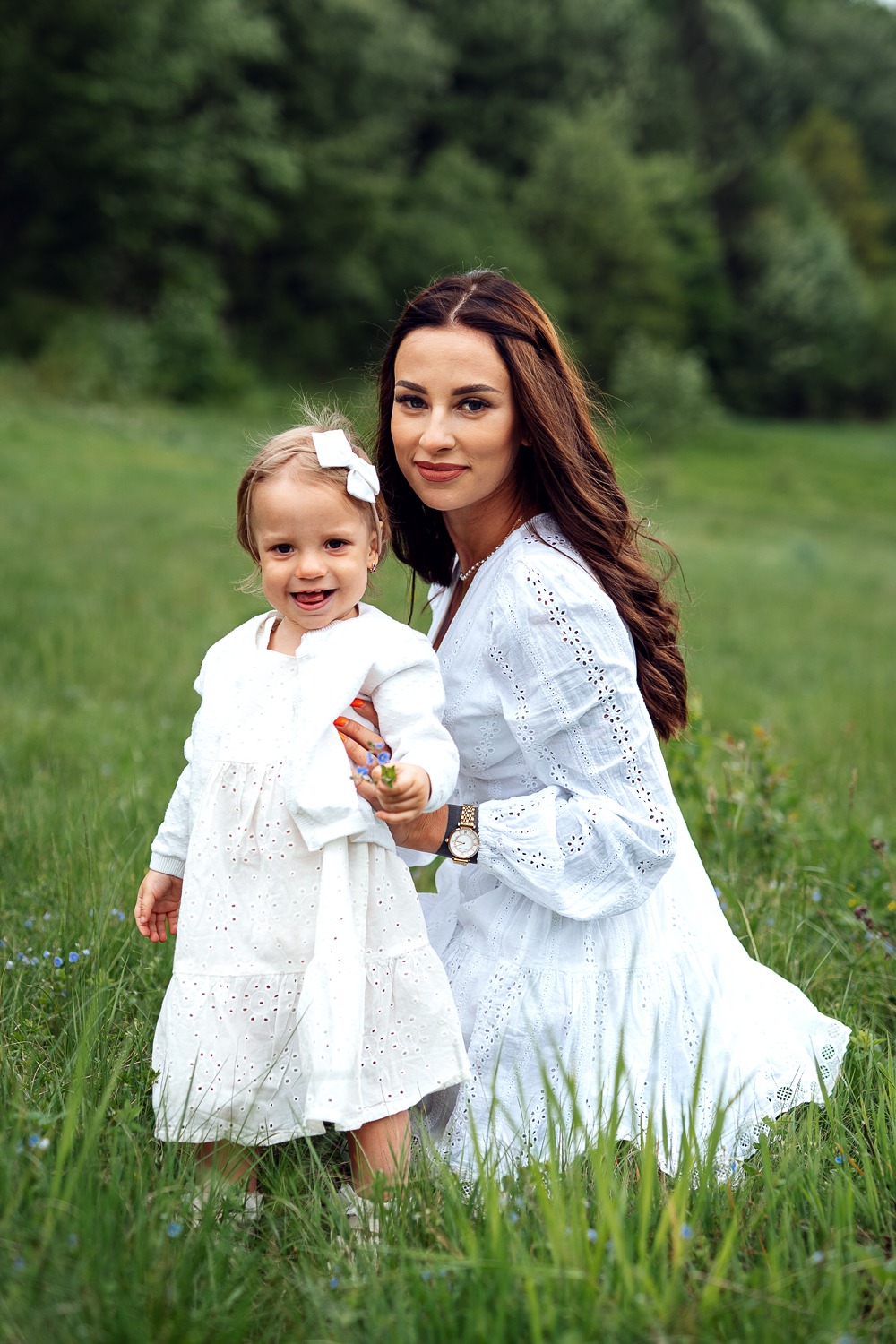 Uśmiechnięta matka z córką w identycznych białych, ażurowych sukienkach, siedząca na łące pełnej trawy i drobnych niebieskich kwiatów, trzymająca mały bukiecik.