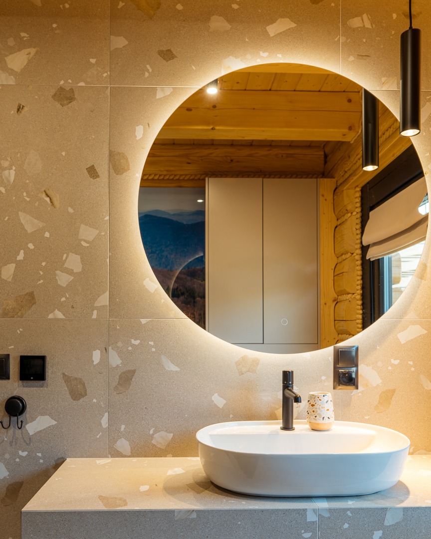 Nowoczesna łazienka z okrągłym lustrem podświetlanym LED, białą umywalką nablatową, czarną armaturą i ścianami wykończonymi płytkami imitującymi lastryko. W odbiciu lustra widoczna drewniana ściana...