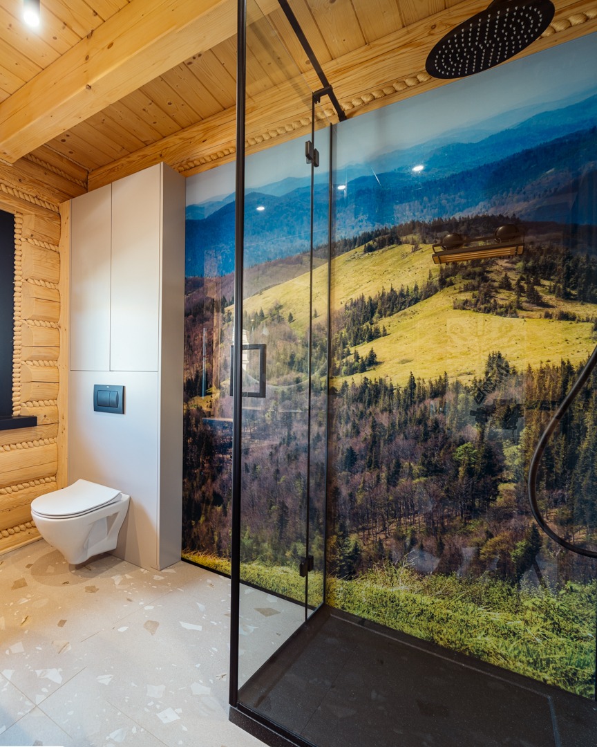 Nowoczesna łazienka z kabiną prysznicową z czarnymi ramami i fototapetą górskiego krajobrazu, drewniane ściany w stylu góralskim, wisząca toaleta i jasna podłoga z lastryko.