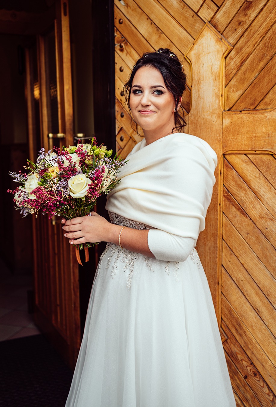 Uśmiechnięta panna młoda w białej sukni z delikatnymi zdobieniami i kremowym szalem, trzymająca bukiet z białych róż i fioletowych kwiatów na tle drewnianych drzwi.