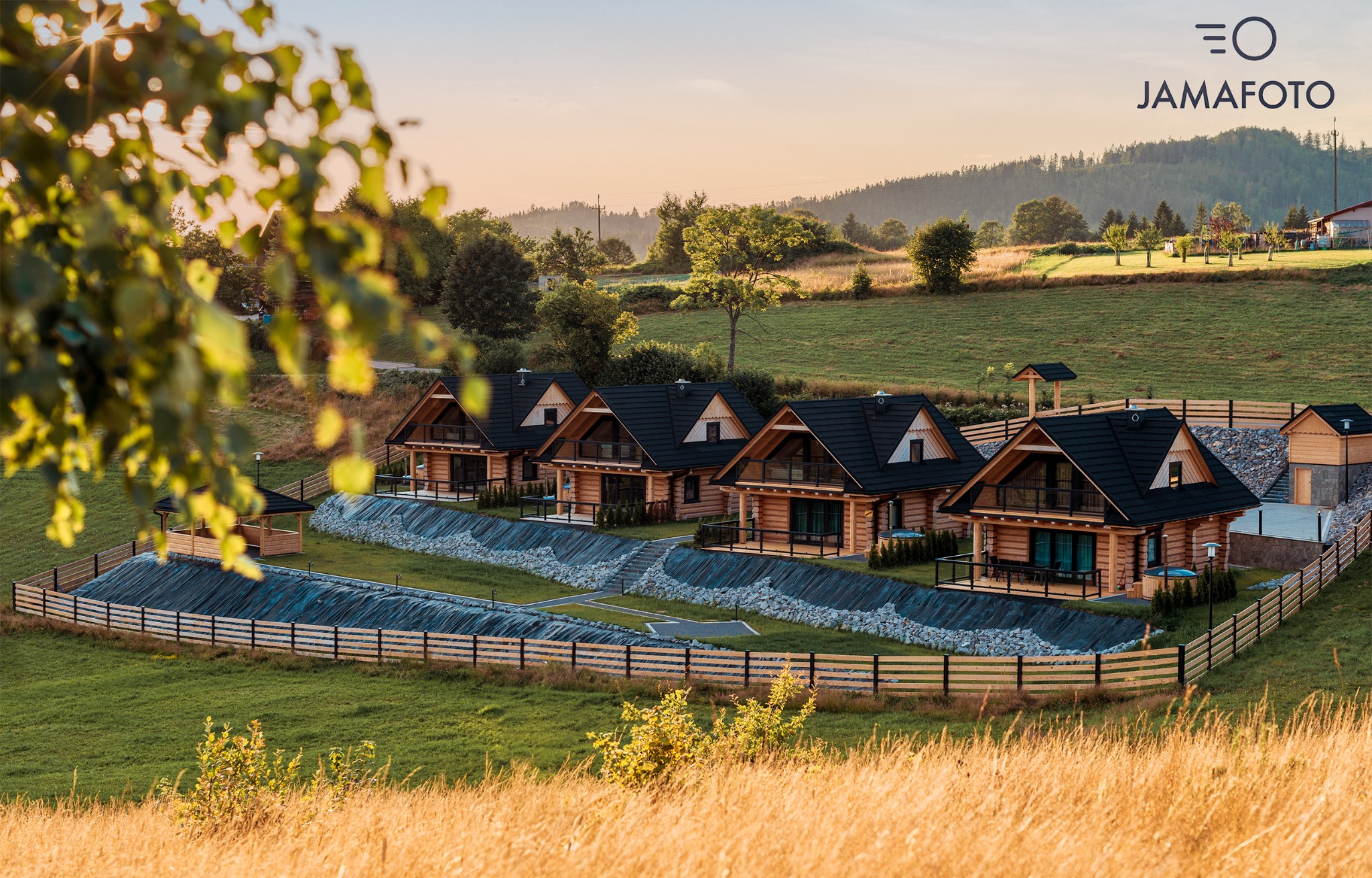 Widok na kompleks drewnianych domków letniskowych z czarnymi dachami, położonych na wzgórzu, z widokiem na góry o zachodzie słońca, sfotografowany z perspektywy trawy na pierwszym planie i z logo...