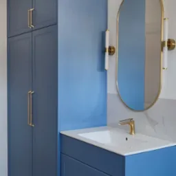 Niebieska szafa łazienkowa na wymiar z wysoką zabudową, złotymi uchwytami i owalnym lustrem podświetlanym kinkietami.