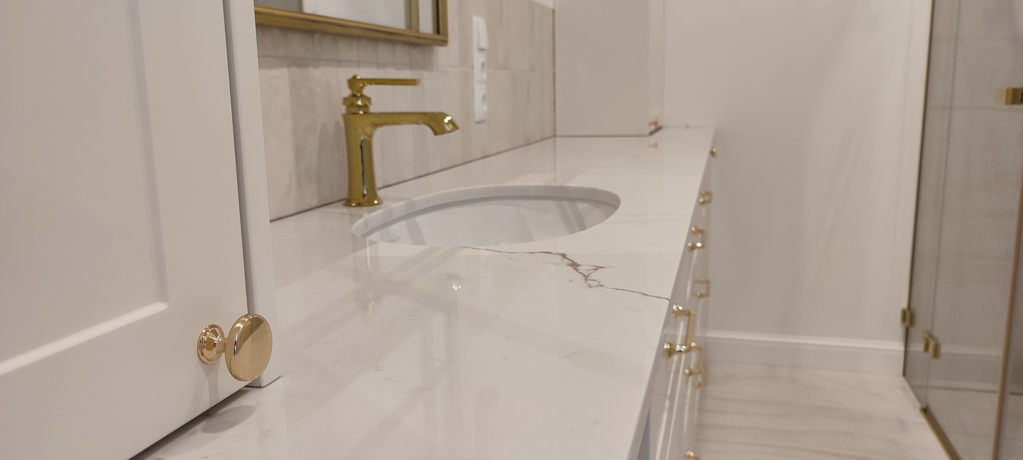 Biała szafka łazienkowa z umywalką wpuszczaną w blat z białego marmuru z złotą armaturą i uchwytami, widok z boku pod kątem.