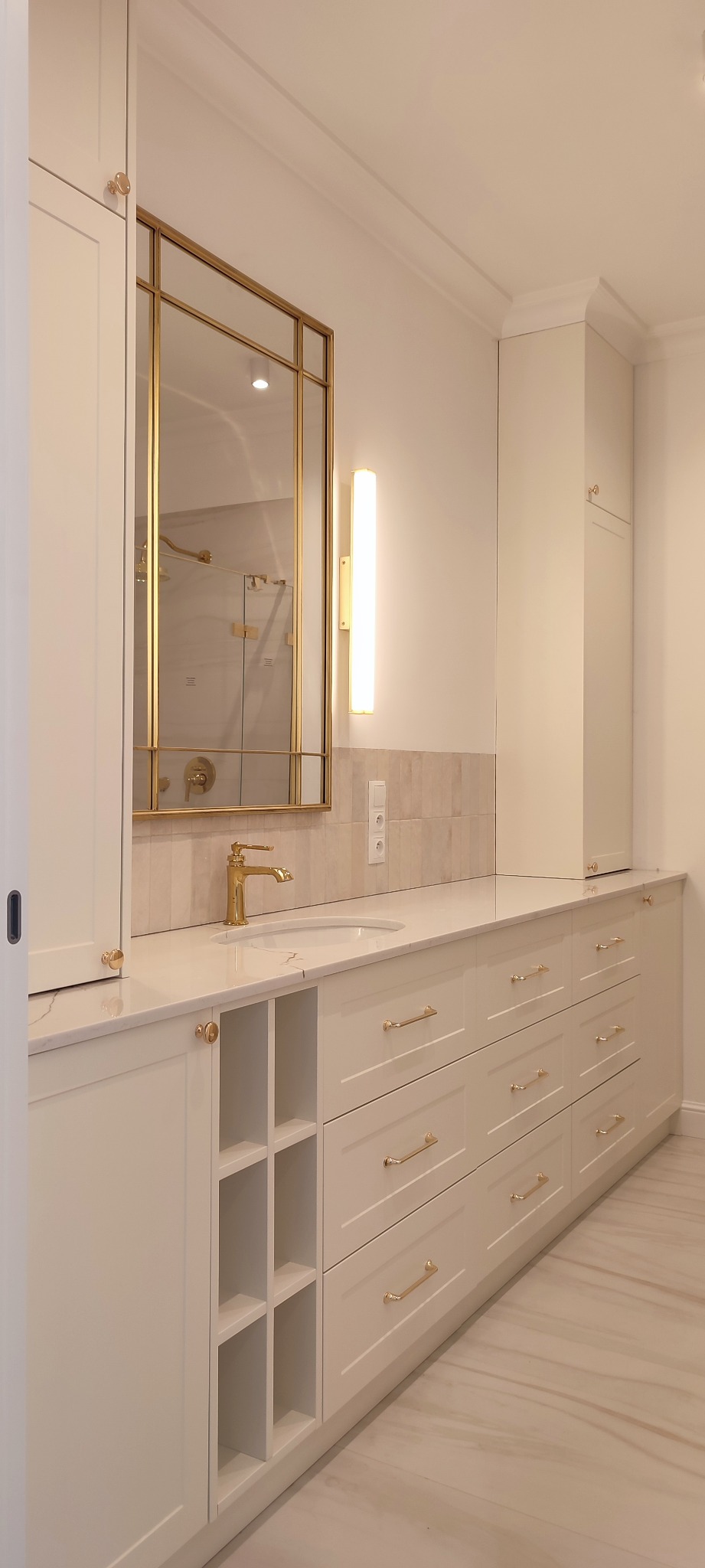 Jasna łazienka z zabudową meblową na wymiar, złota armatura i lustro w złotej ramie, widoczna część kabiny prysznicowej w odbiciu.