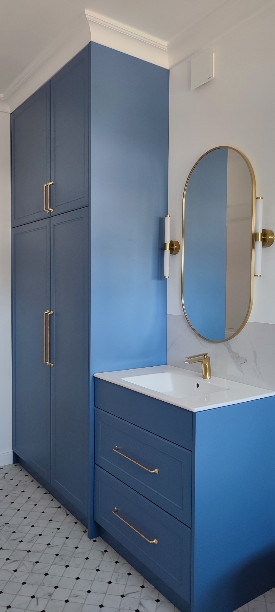 Niebieska szafa łazienkowa na wymiar z wysoką zabudową, złotymi uchwytami i owalnym lustrem podświetlanym kinkietami.