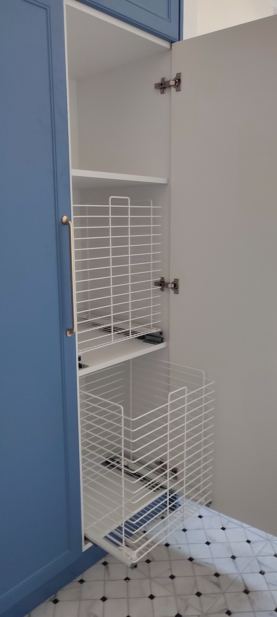 Wnętrze szafy z niebieskimi drzwiami, wyposażonej w wysuwane, białe, metalowe kosze na prowadnicach, widok na marmurową podłogę.