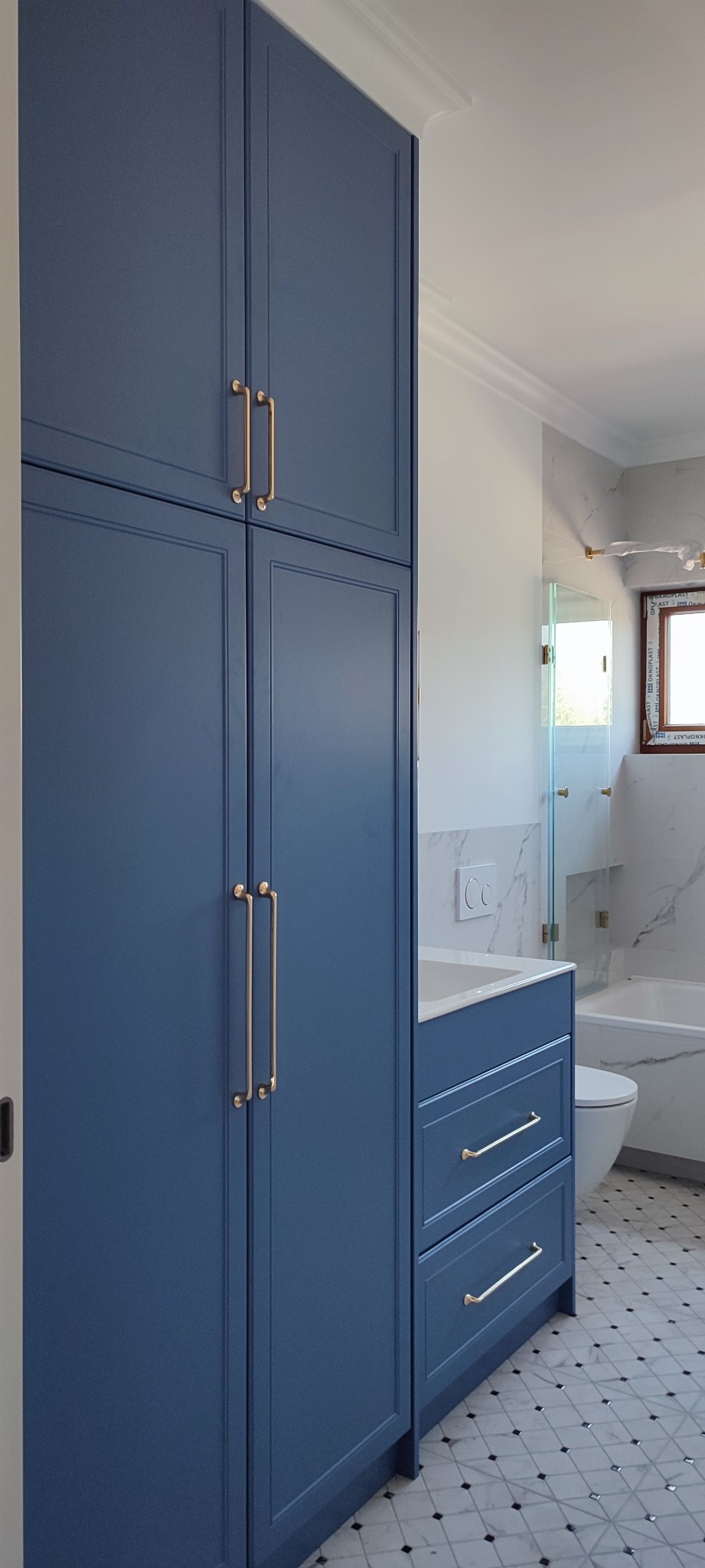 Niebieska zabudowa meblowa w łazience z wysoką szafą i szufladami, złote uchwyty, marmurowe wykończenie, mozaikowa podłoga.