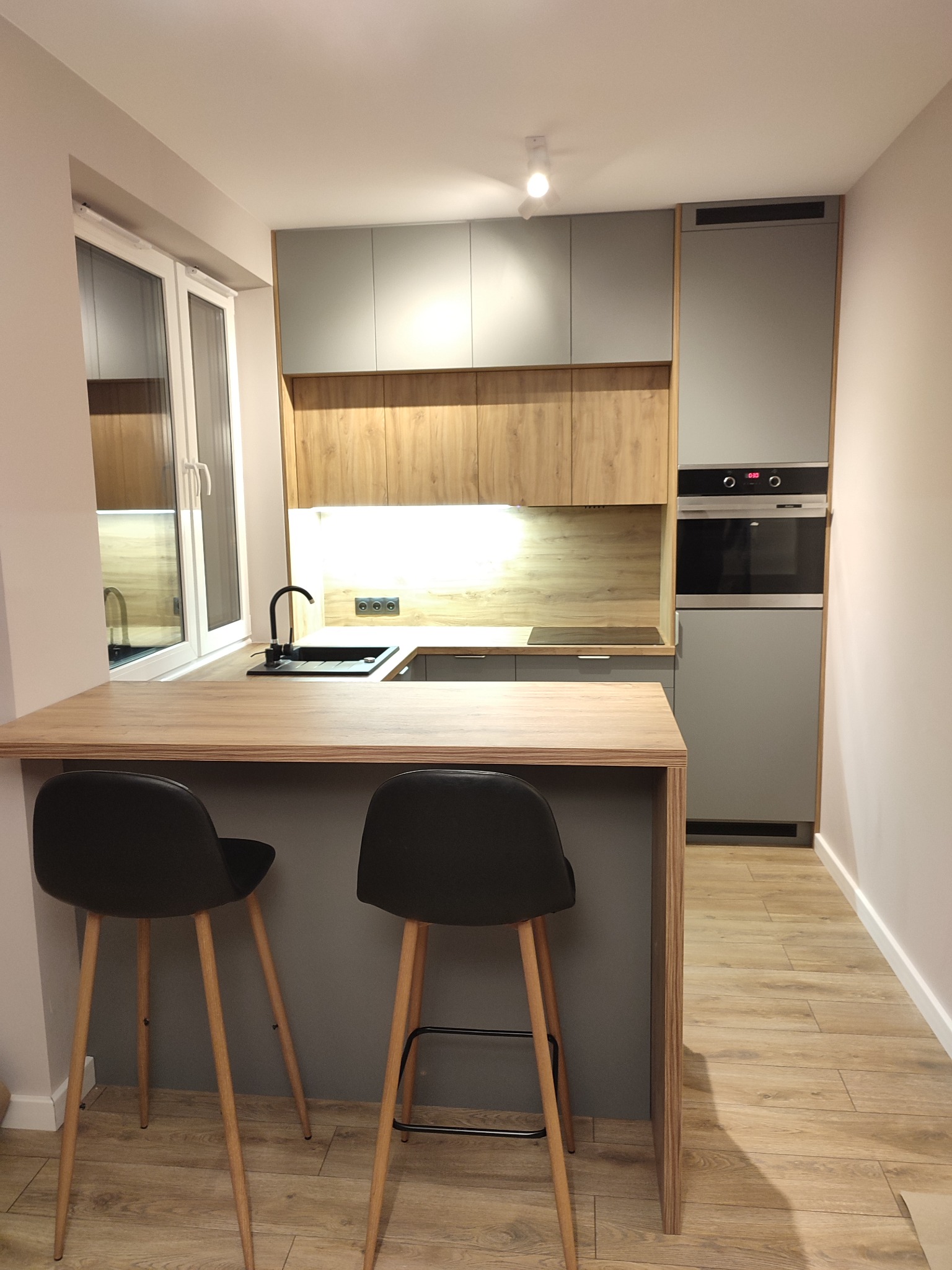 Nowoczesna kuchnia z szarymi szafkami i drewnianym blatem, wyspą z dwoma czarnymi hokerami na drewnianych nogach, oświetleniem podszafkowym i widocznym piekarnikiem w zabudowie.