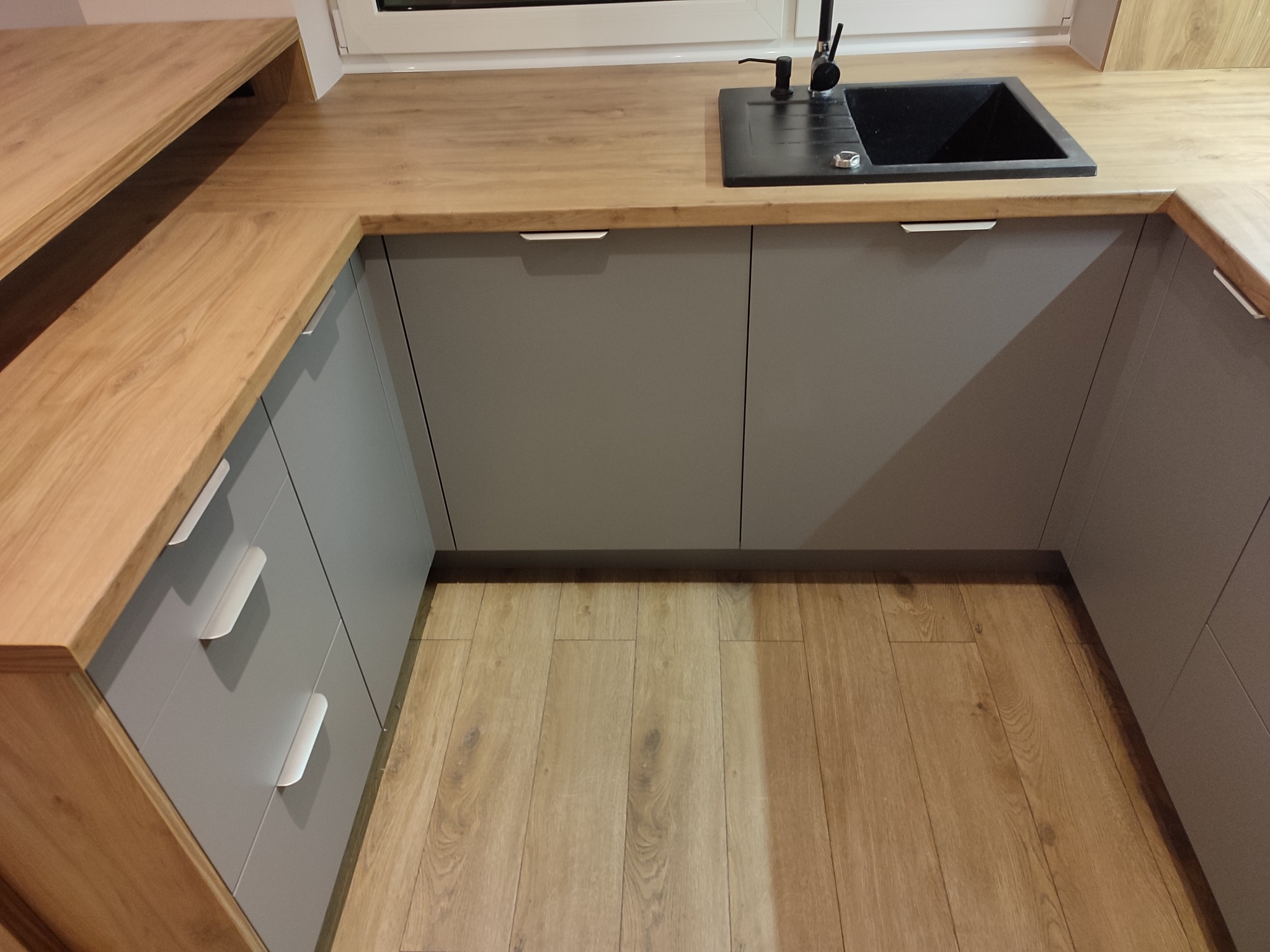 Szare, matowe szafki kuchenne z drewnianym blatem i czarnym, kwadratowym zlewem, widok z góry na drewnianą podłogę.