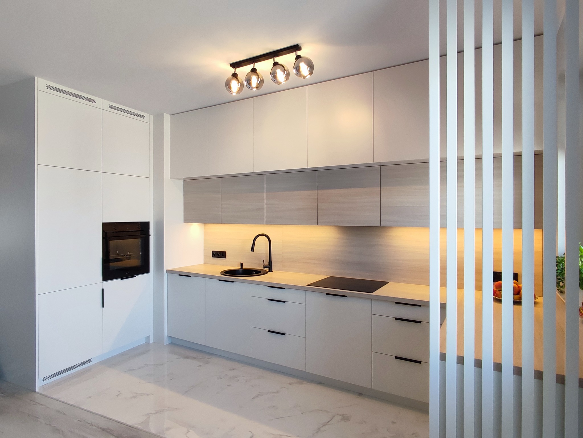 Nowoczesna kuchnia z białymi szafkami, drewnopodobnym blatem i czarną armaturą, oświetlona lampą sufitową z kulistymi kloszami, oddzielona od reszty pomieszczenia ażurową ścianką z białych listew.