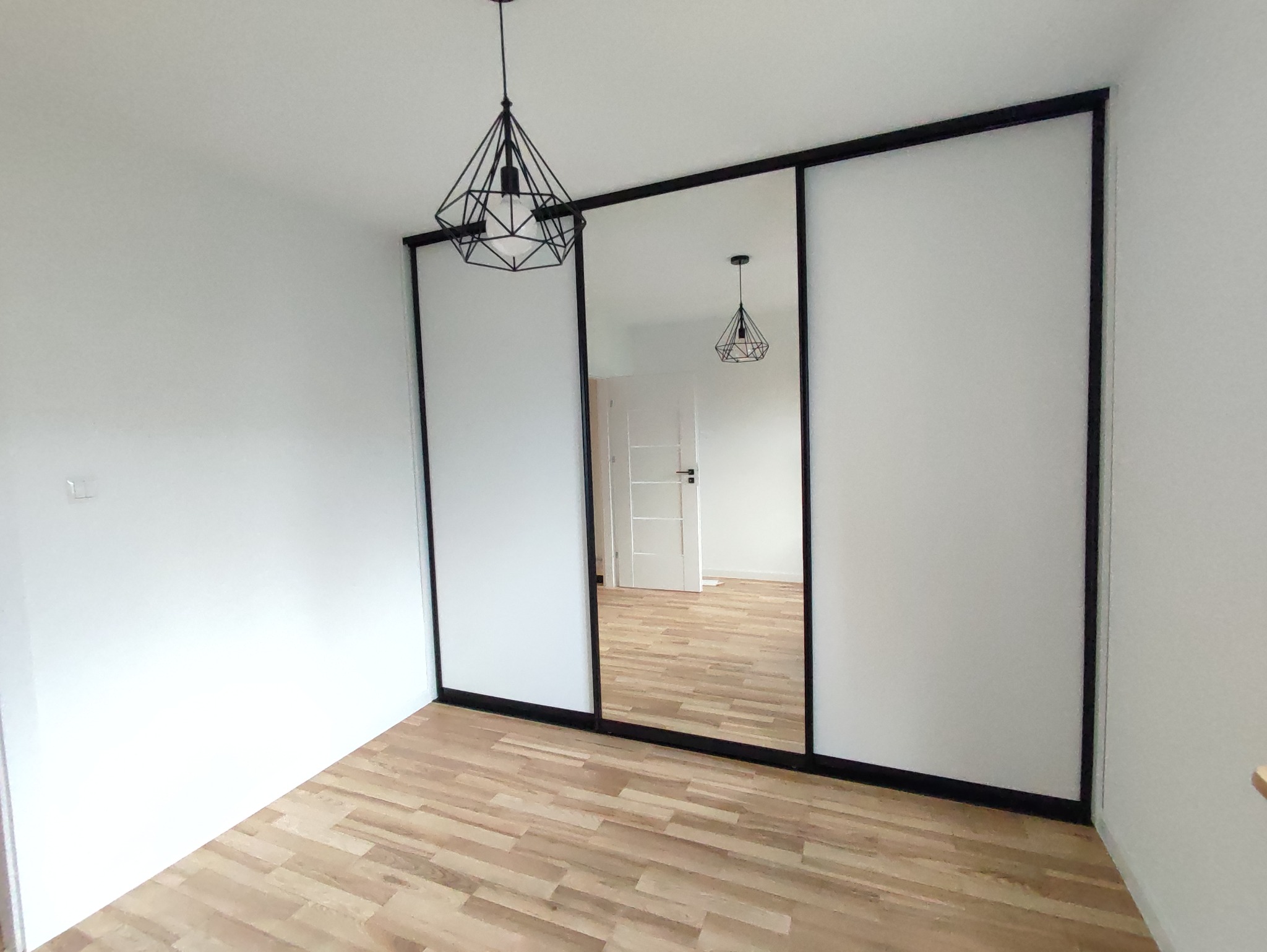 Nowoczesna szafa przesuwna z czarnymi ramami i lustrzanym panelem w minimalistycznym wnętrzu z drewnianą podłogą i geometryczną lampą sufitową.