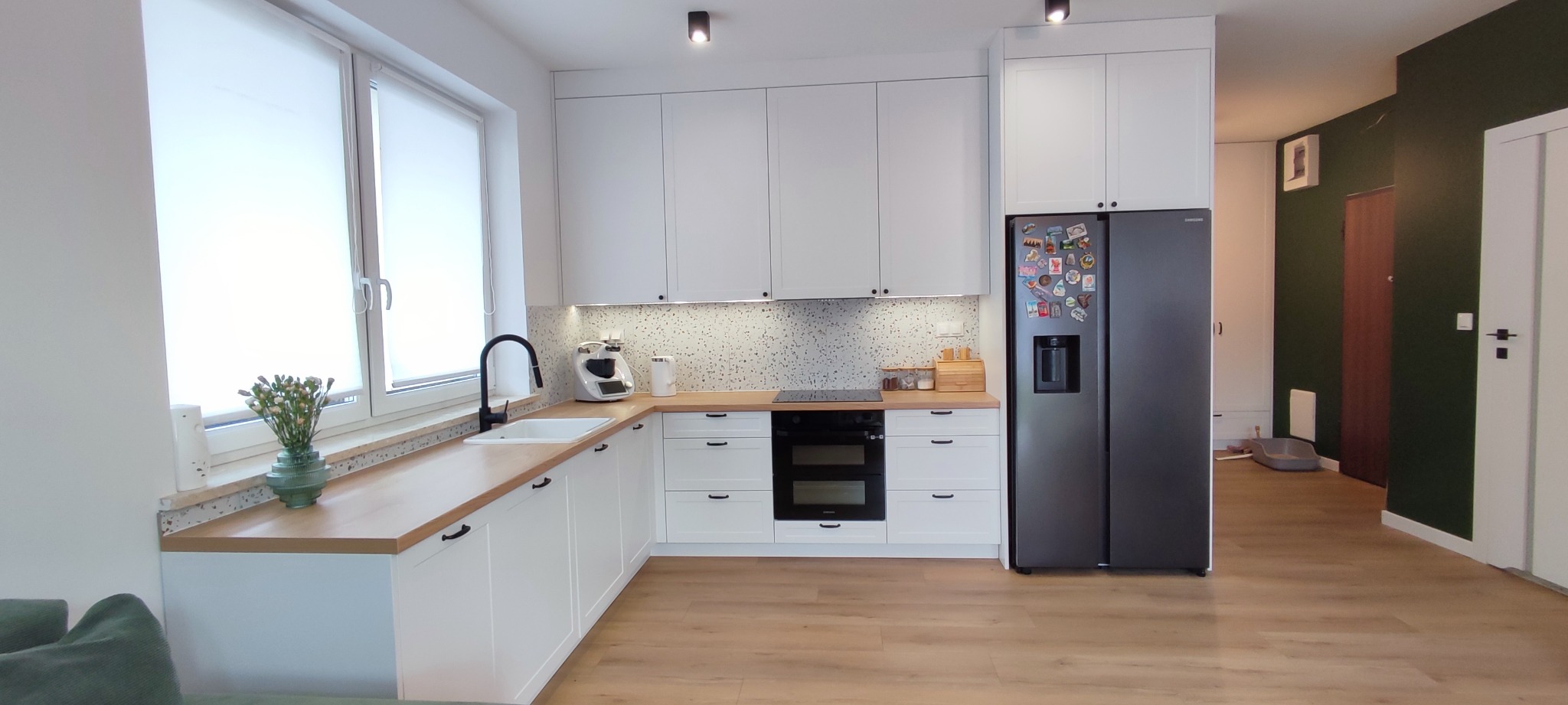 Jasna kuchnia z białymi szafkami i drewnianym blatem, czarny zlew i bateria przy oknie, na blacie kwiaty w zielonym wazonie, w tle ciemnozielona ściana w przedpokoju.