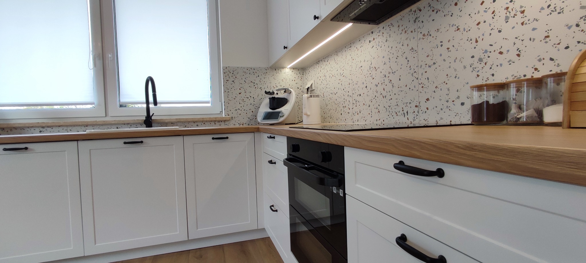 Jasna kuchnia z białymi szafkami, drewnianym blatem i mozaikową ścianą nad blatem. Widoczny piekarnik, zlew z czarną armaturą oraz akcesoria kuchenne na blacie.