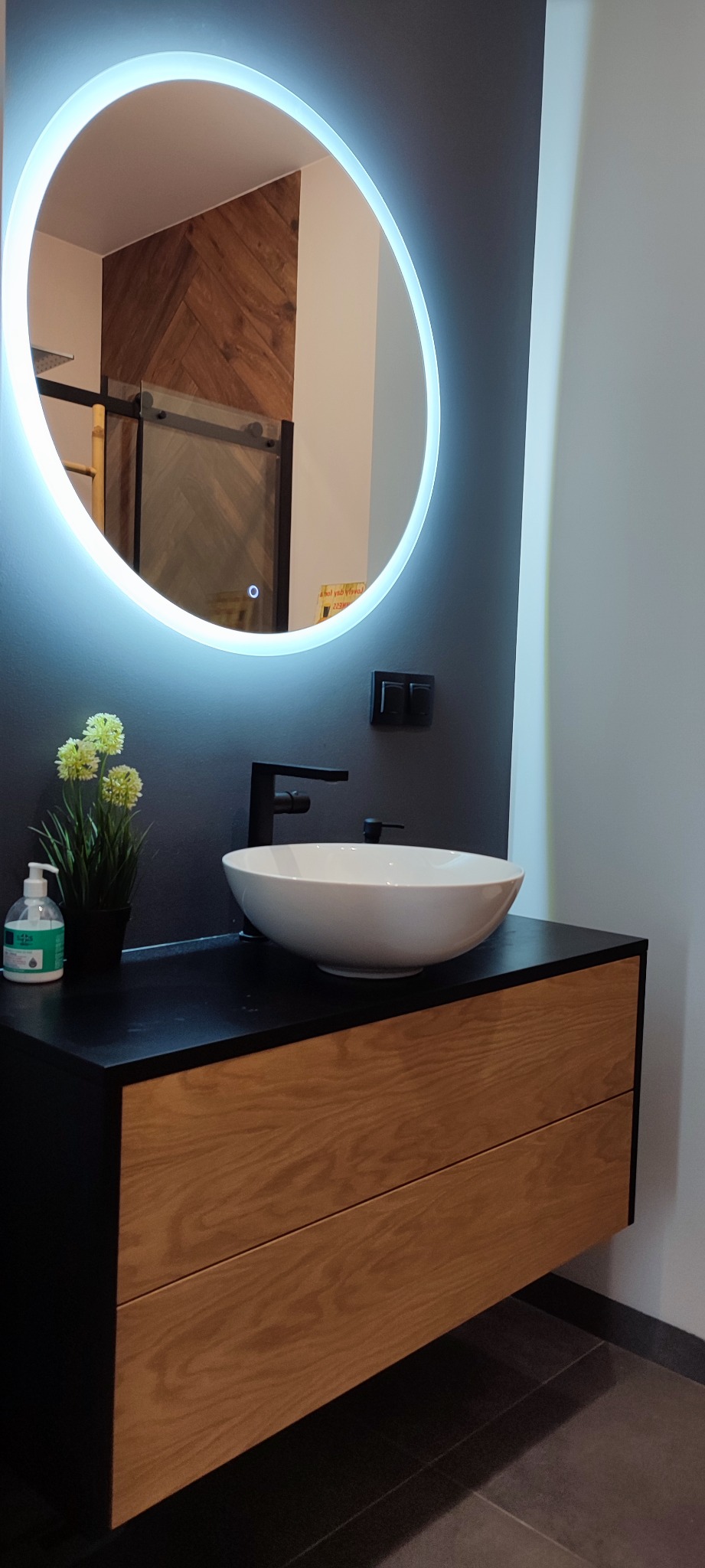 Nowoczesna łazienka z okrągłym lustrem podświetlanym LED, umywalką nablatową i szafką z drewnianymi frontami. W tle widoczna ściana z drewnianą okładziną.