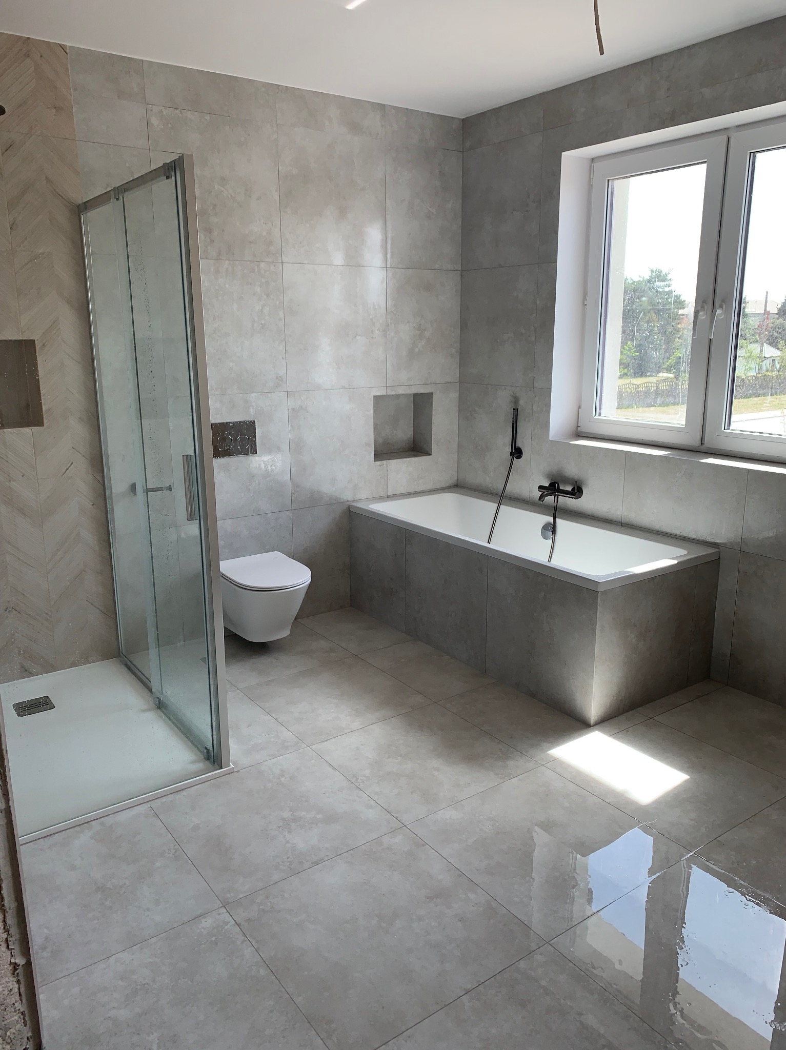 Nowoczesna łazienka w odcieniach szarości z kabiną prysznicową typu walk-in, podwieszaną toaletą i wanną pod oknem, wykończona płytkami imitującymi kamień, z widocznymi elementami armatury...