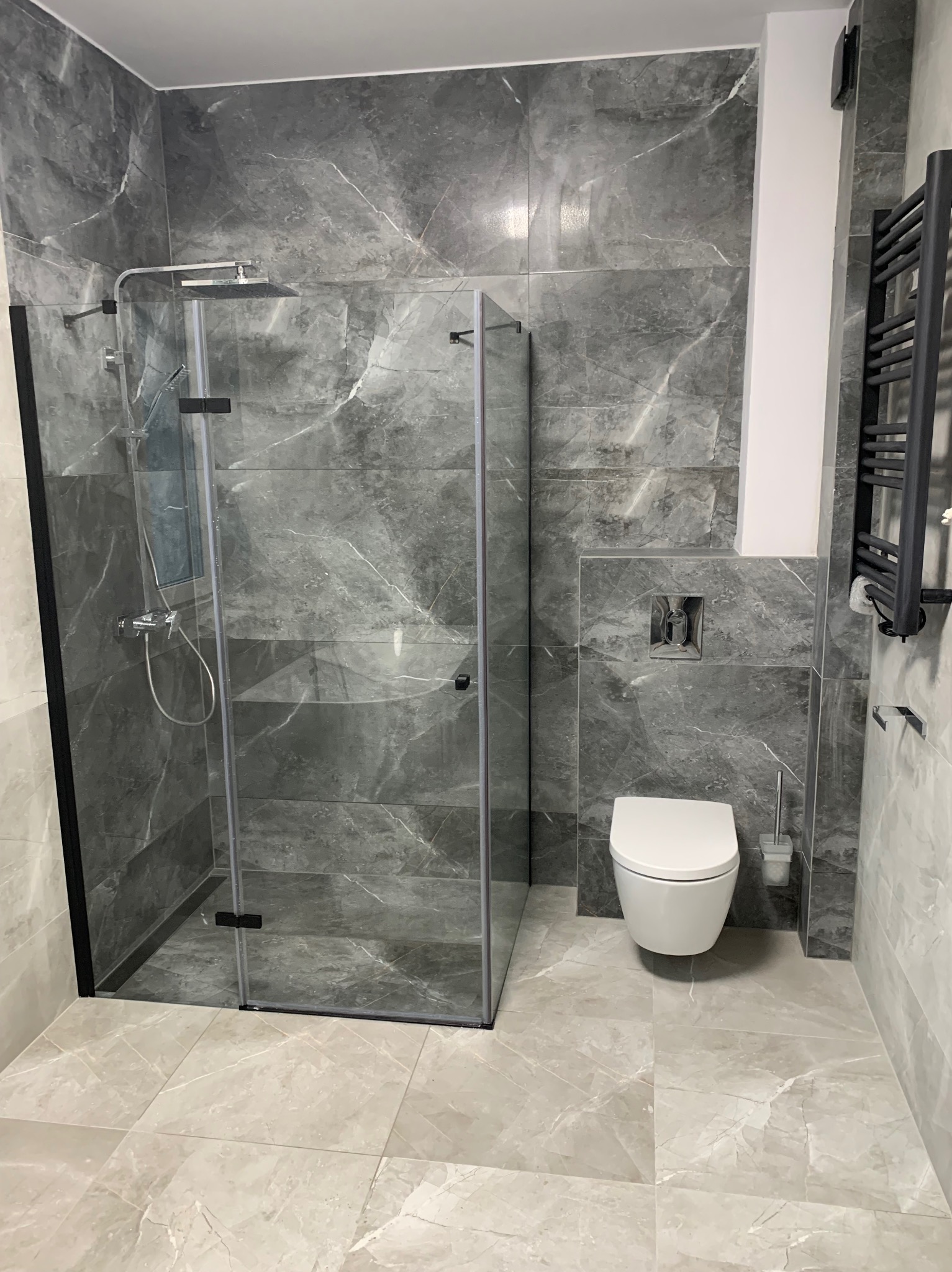 Nowoczesna łazienka z kabiną prysznicową typu walk-in, wiszącą toaletą i grzejnikiem drabinkowym w odcieniach szarości z marmurkowym wzorem na płytkach.