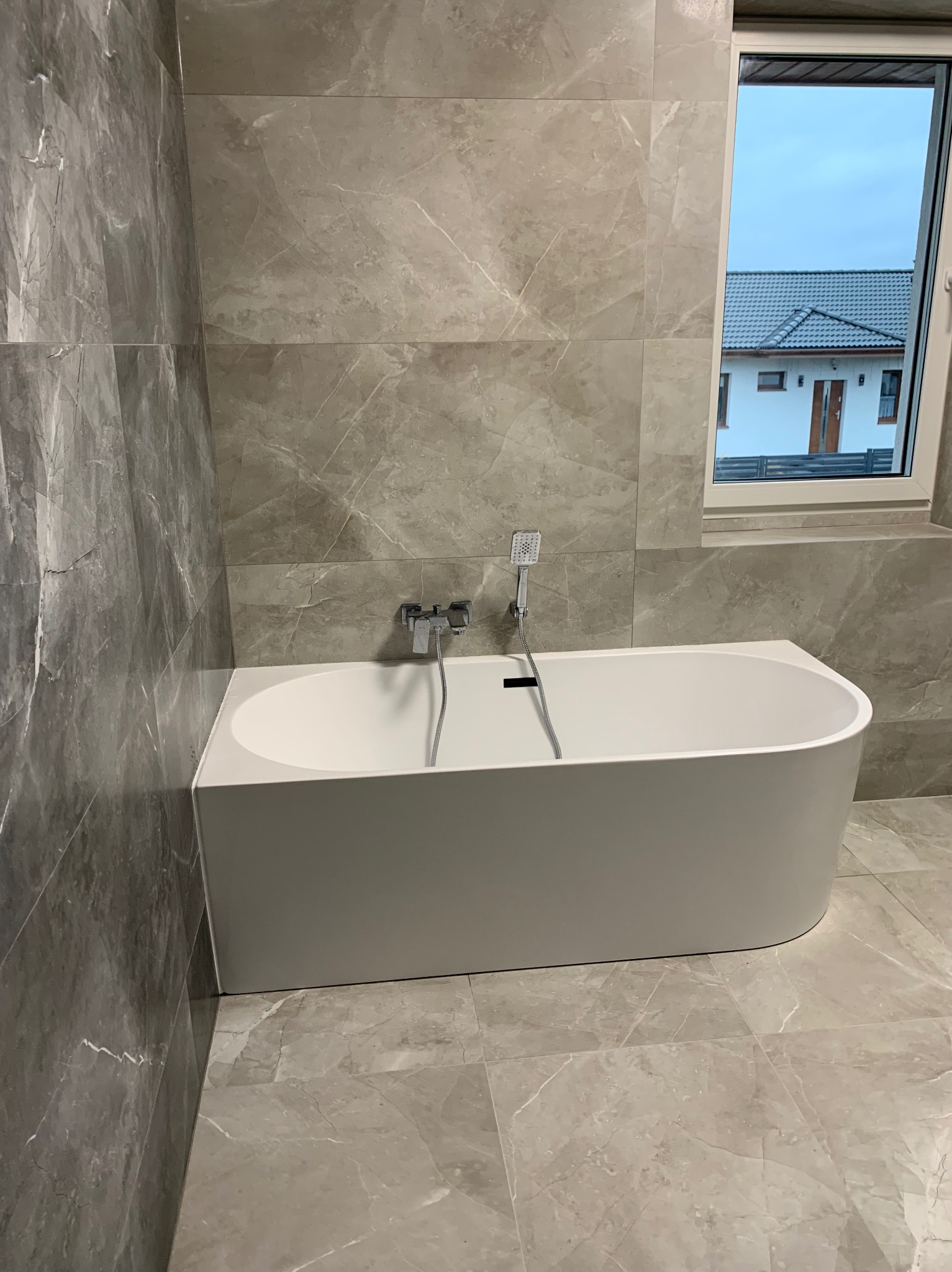 Nowoczesna, wolnostojąca wanna w łazience wykończonej płytkami imitującymi marmur, z widokiem na dom przez okno.